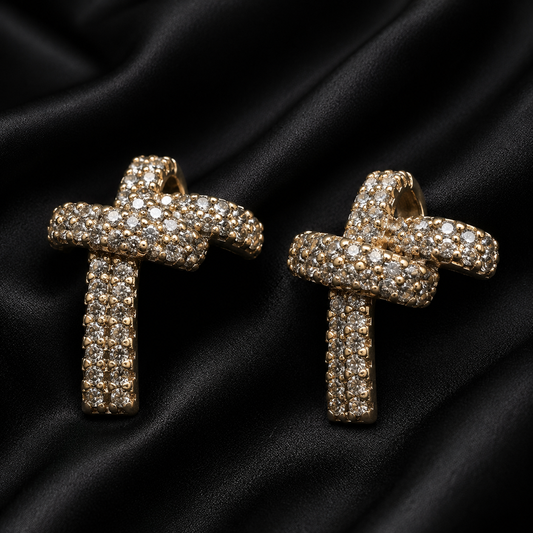 A pair of gold cross-shaped earrings covered in small clear stones, displayed on a black satin fabric background.