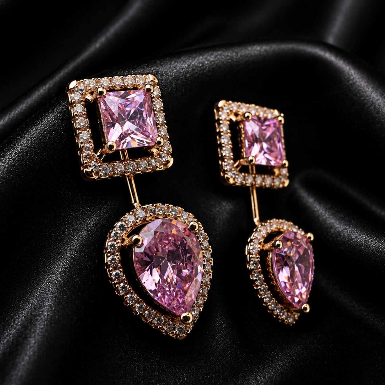 A pair of gold-tone drop earrings featuring a square pink gemstone at the top and a larger pear-shaped pink gemstone at the bottom, both surrounded by small clear stones, displayed on a black satin fabric background.