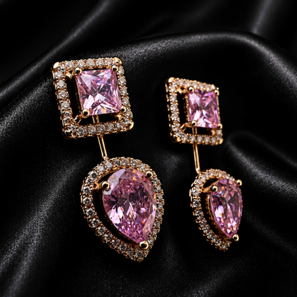 A pair of gold-tone drop earrings featuring a square pink gemstone at the top and a larger pear-shaped pink gemstone at the bottom, both surrounded by small clear stones, displayed on a black satin fabric background.