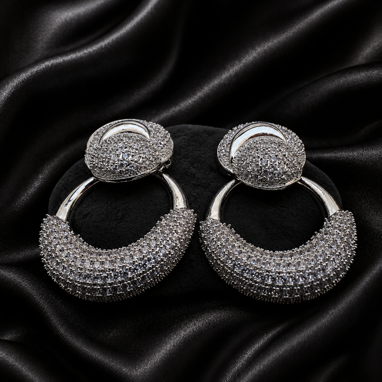 A pair of silver-toned hoop earrings covered in clear pavé stones, displayed on a black satin fabric background.