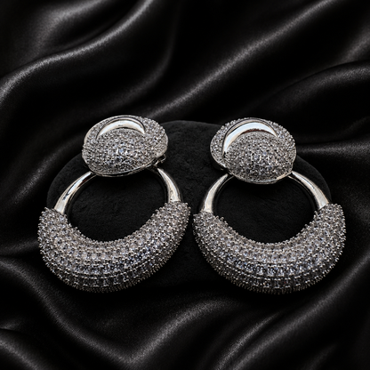 A pair of silver-toned hoop earrings covered in clear pavé stones, displayed on a black satin fabric background.