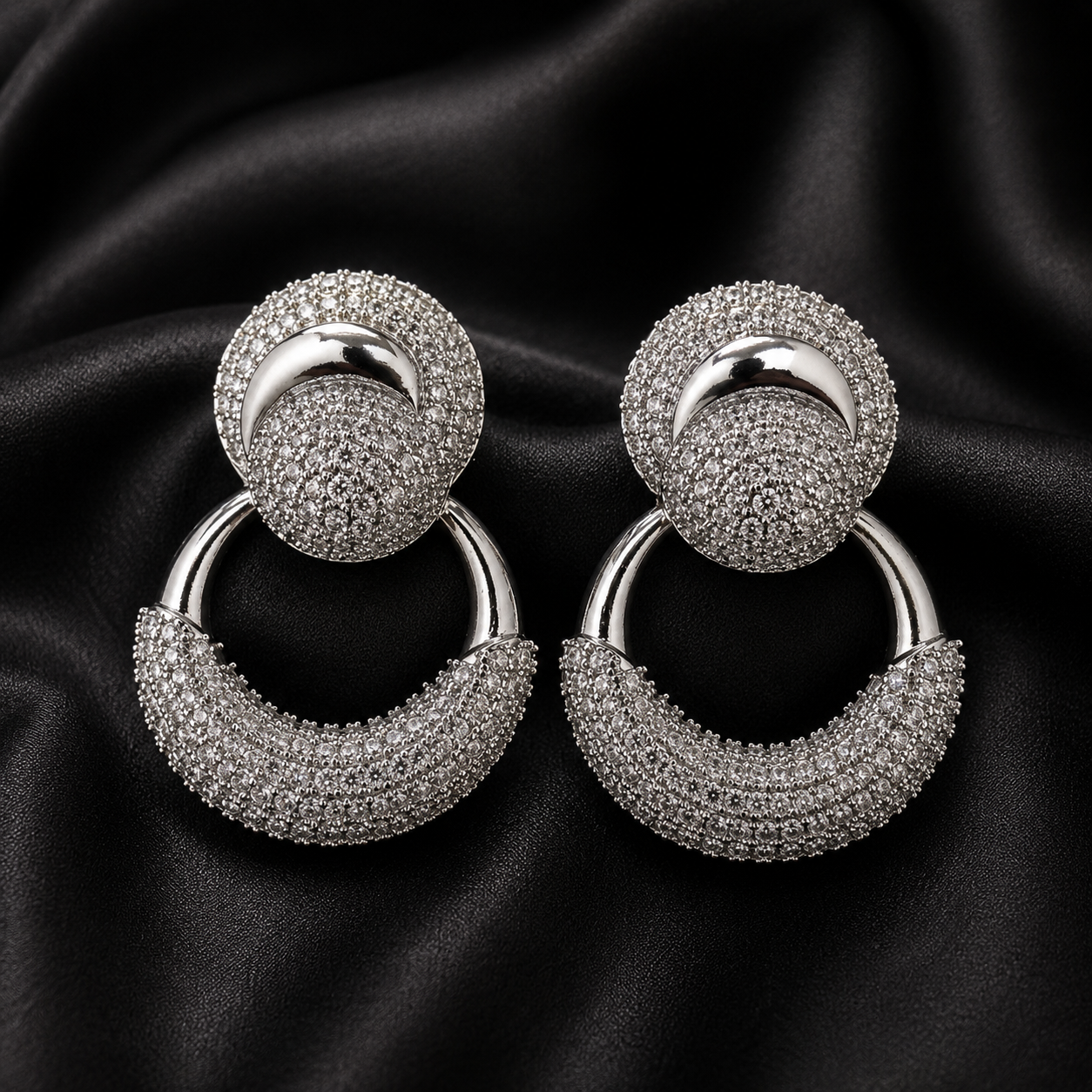 A pair of silver-toned drop hoop earrings with circular studs covered in small clear stones, displayed on a black satin fabric background.
