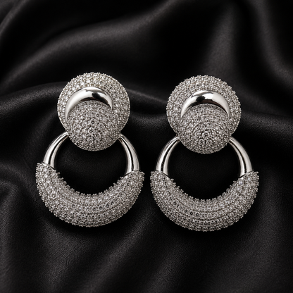 A pair of silver-toned drop hoop earrings with circular studs covered in small clear stones, displayed on a black satin fabric background.