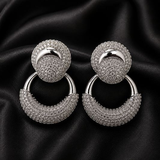 A pair of silver-toned drop hoop earrings with circular studs covered in small clear stones, displayed on a black satin fabric background.