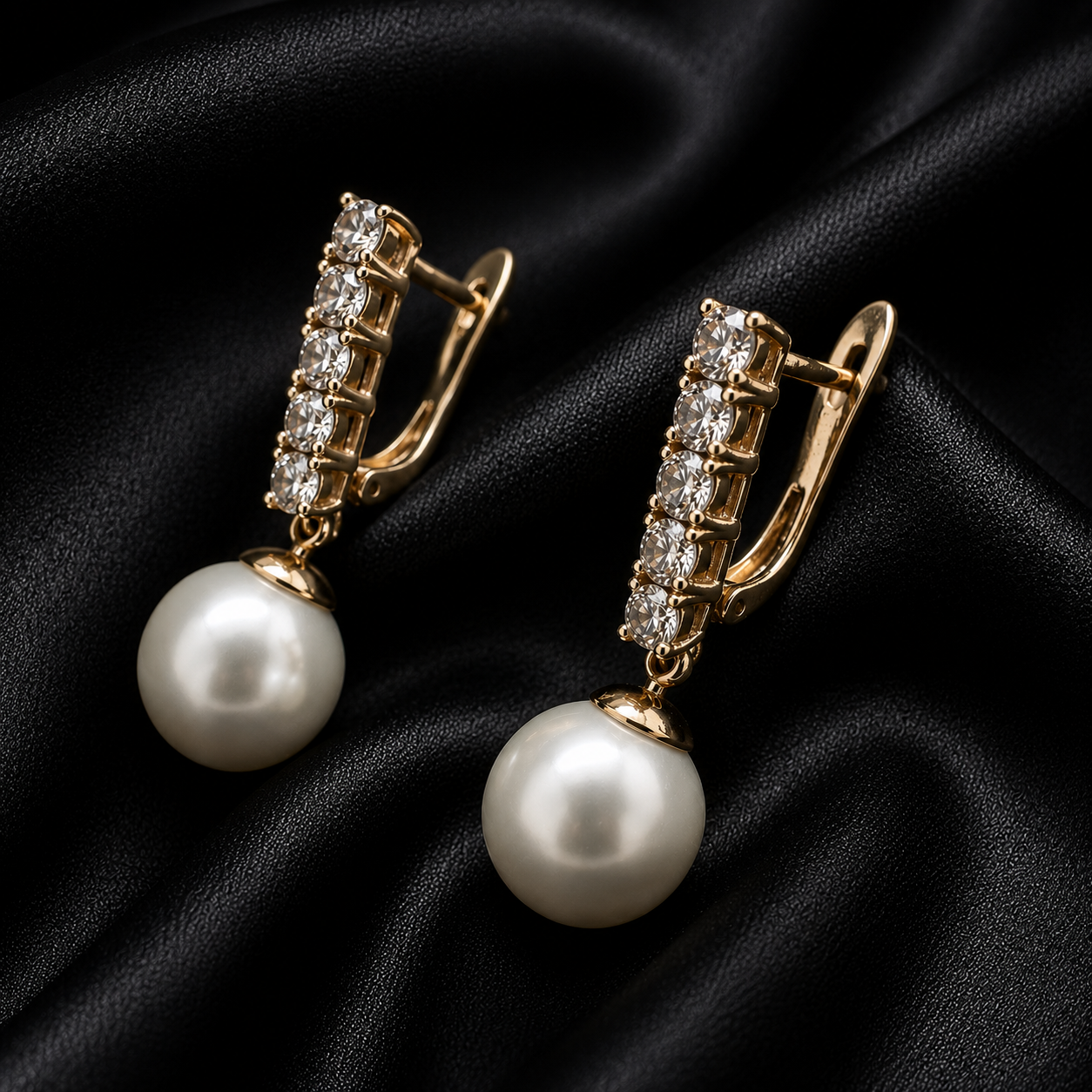 A pair of gold-toned drop earrings with a row of small clear stones and a large white pearl dangle, displayed on a black satin fabric background.