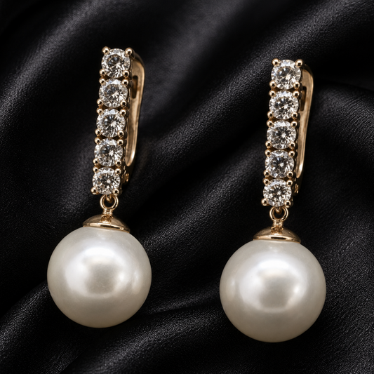 A pair of gold drop earrings with a row of clear cubic zirconia stones and a large white pearl dangling below, displayed on a black satin fabric background.