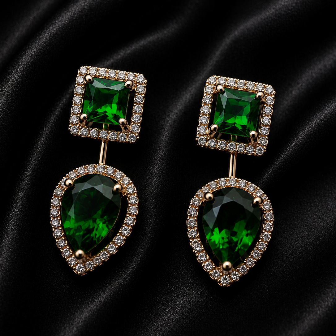 A pair of gold drop earrings featuring a square-cut green gemstone in a halo setting on top and a larger teardrop-shaped green gemstone in a halo setting below, displayed on a black satin background.