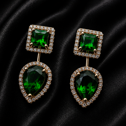 A pair of gold drop earrings featuring a square-cut green gemstone in a halo setting on top and a larger teardrop-shaped green gemstone in a halo setting below, displayed on a black satin background.