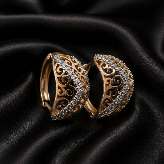 A pair of gold crescent-shaped hoop earrings with intricate scrollwork filigree and rows of clear crystals, displayed on a draped black satin fabric background.