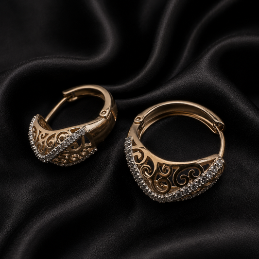 A pair of gold-toned hoop earrings with intricate filigree cutouts and rows of small clear stones along the front, displayed on draped black satin fabric.