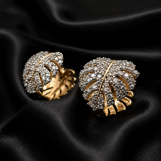 A pair of gold-toned leaf-shaped earrings covered with clear crystals, displayed on a draped black satin fabric background.