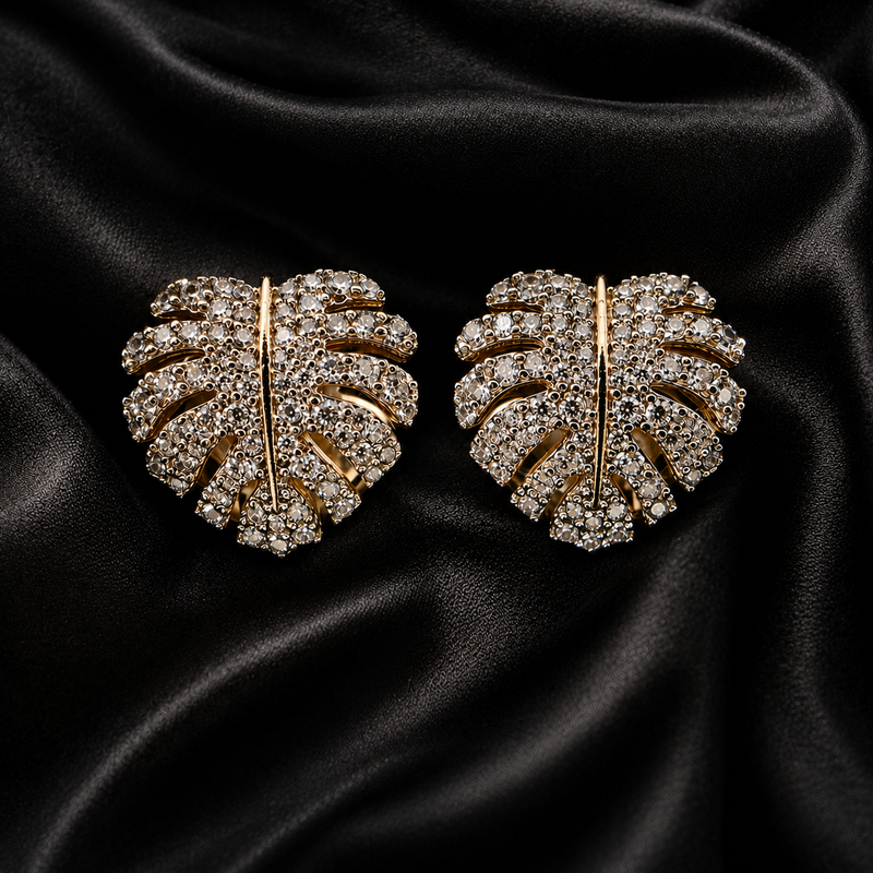 A pair of gold-toned leaf-shaped stud earrings paved with clear stones, displayed on a draped black satin fabric background.