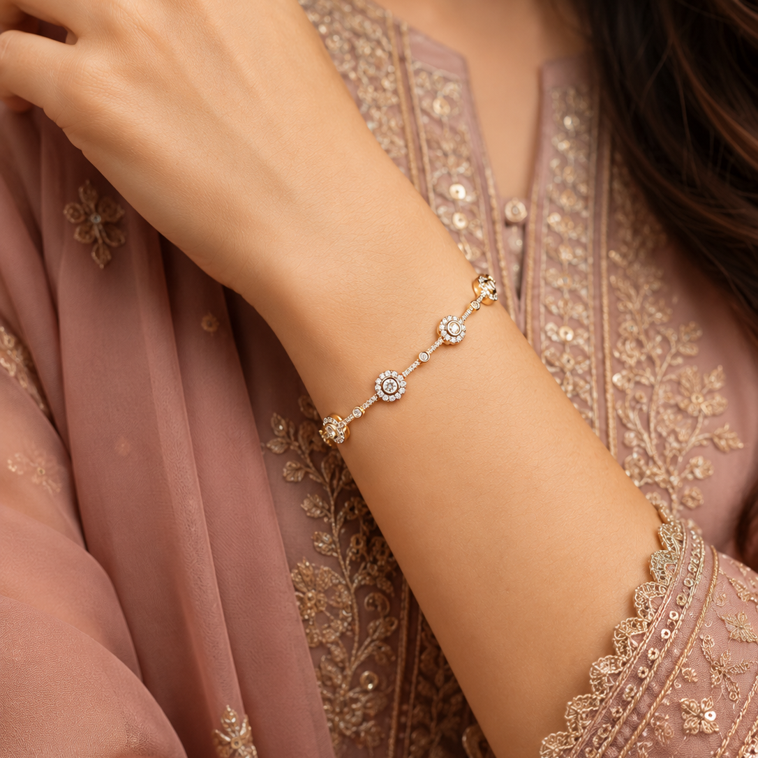 A delicate gold-toned chain bracelet with three central floral motifs, worn on a woman's wrist. The woman is wearing a dusty rose embroidered traditional outfit.