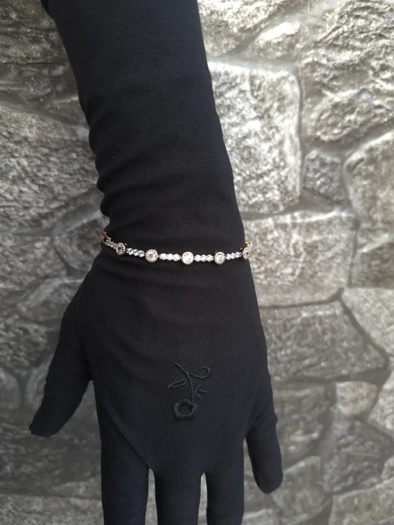 Hand wearing a black glove with a bracelet against a stone wall background
