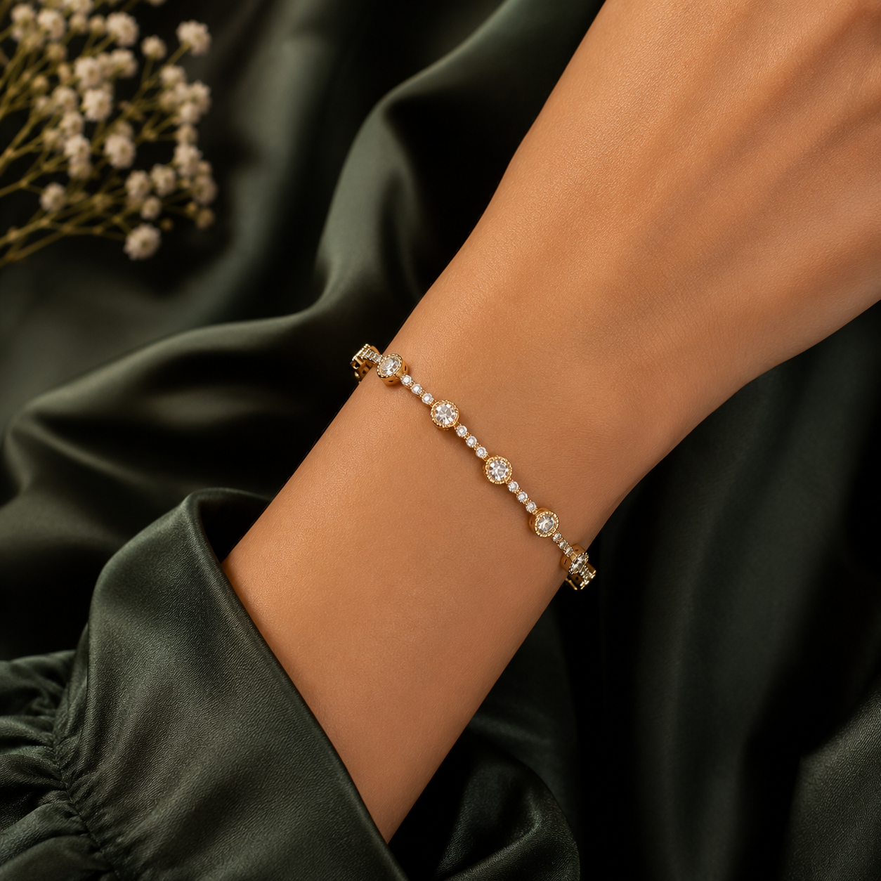 A delicate gold bracelet with small clear gemstones and pearl-like accents, worn on a woman's wrist against a dark green satin fabric background.
