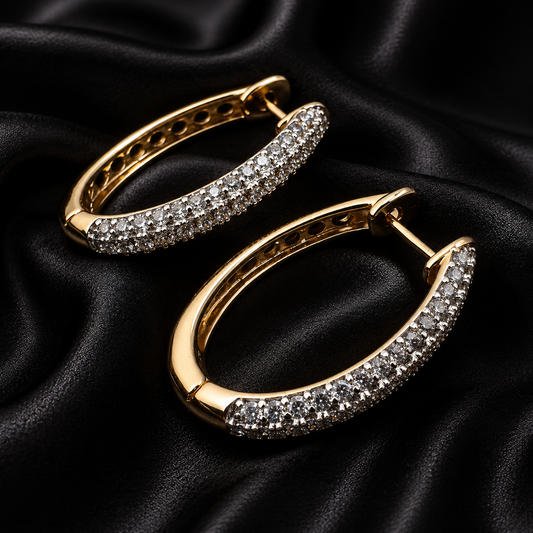 A pair of gold hoop earrings paved with small clear stones, displayed on a draped black satin fabric background.