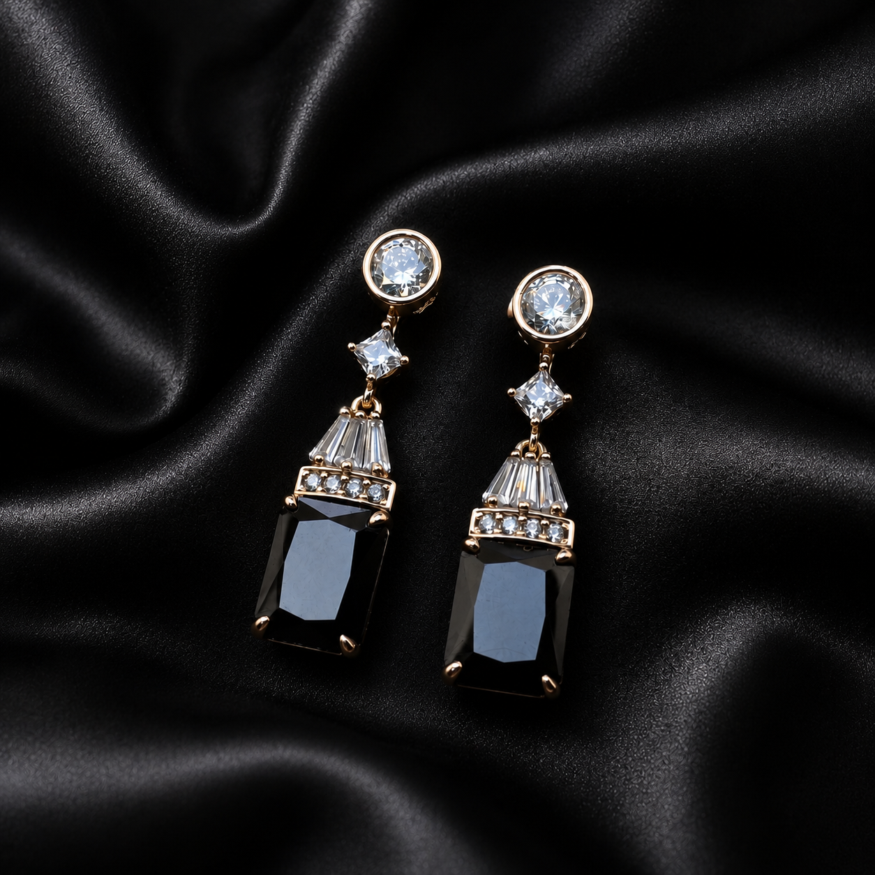 A pair of gold-tone drop earrings with clear round and baguette crystals, featuring rectangular black gemstone drops, displayed on a draped black satin fabric background.