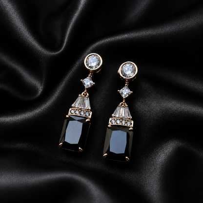 A pair of gold-tone drop earrings with clear round and baguette crystals, featuring rectangular black gemstone drops, displayed on a draped black satin fabric background.