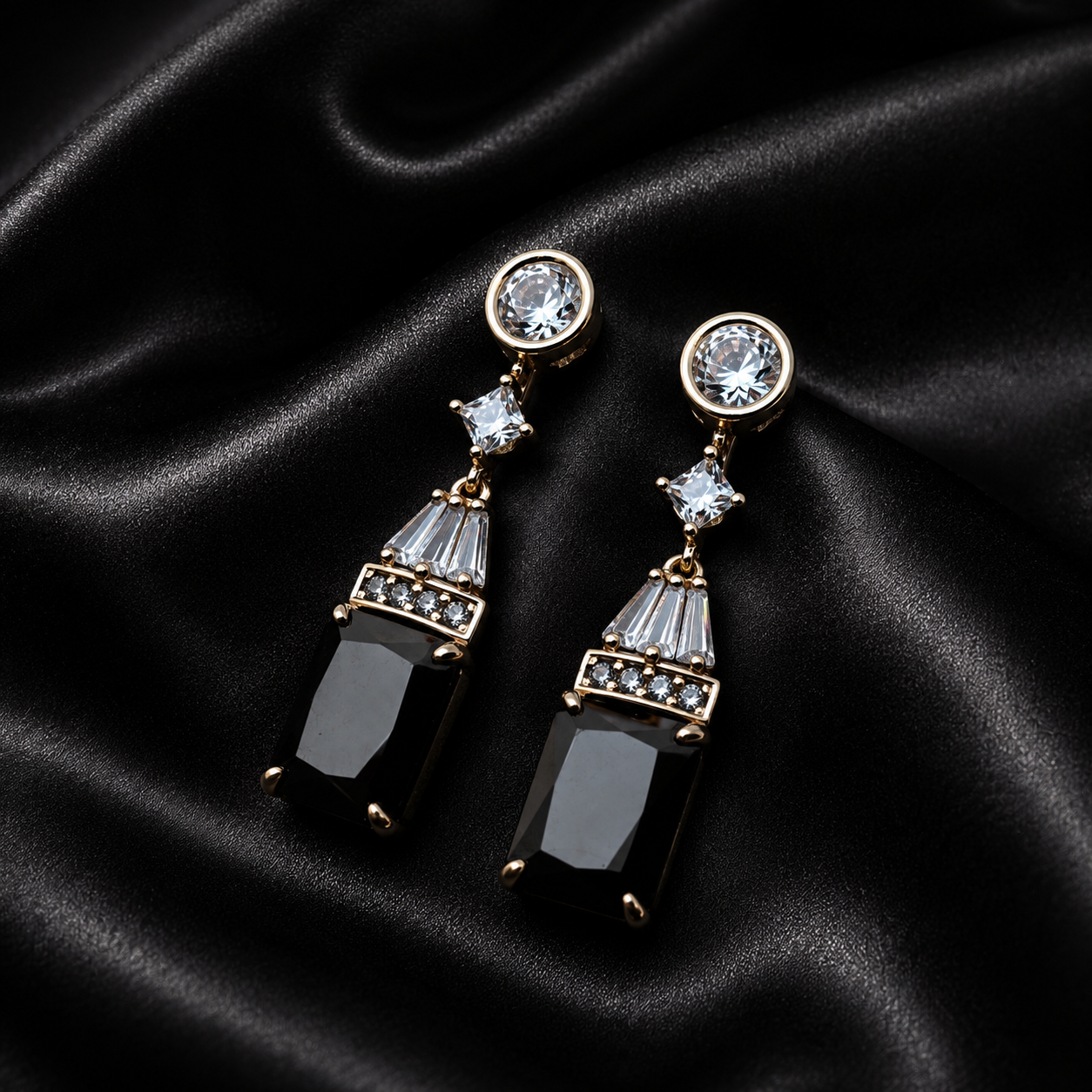 A pair of gold-tone drop earrings with clear round stud tops, small clear crystal accents, and large black rectangular drop pendants, displayed on a black satin fabric background.