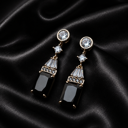 A pair of gold-tone drop earrings with clear round stud tops, small clear crystal accents, and large black rectangular drop pendants, displayed on a black satin fabric background.