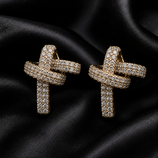 A pair of gold-toned cross-shaped earrings paved with small clear stones, displayed on a draped black satin fabric background.