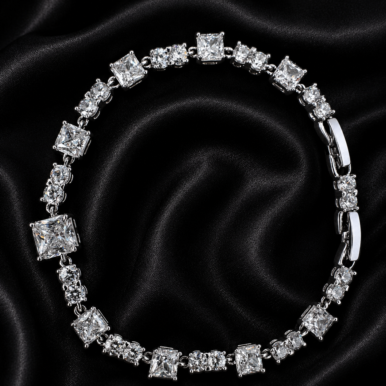 A silver-toned tennis bracelet featuring princess-cut clear stones, displayed on a black satin fabric background.