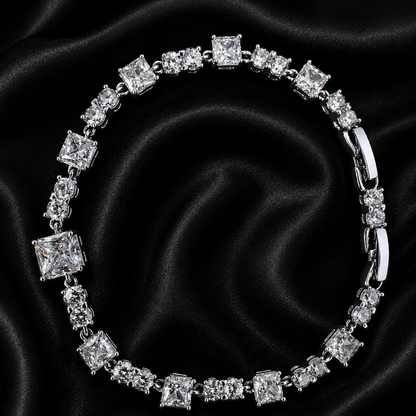A silver-toned tennis bracelet featuring princess-cut clear stones, displayed on a black satin fabric background.