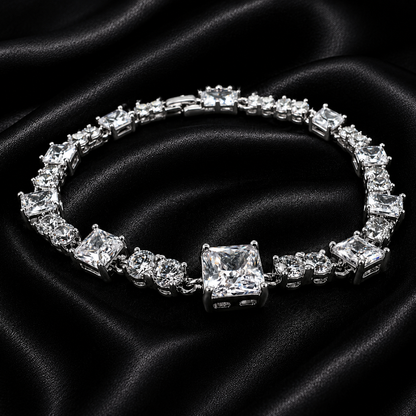 A silver-toned tennis bracelet featuring a large princess-cut clear center stone and multiple clear rectangular and round accent stones, displayed on a draped black satin fabric background.