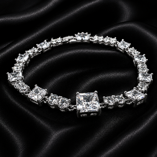 A silver-toned tennis bracelet featuring a large princess-cut clear center stone and multiple clear rectangular and round accent stones, displayed on a draped black satin fabric background.