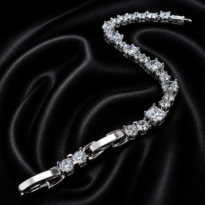 A silver tennis bracelet with clear round-cut stones, arranged in a curved S-shape on a black satin fabric background.