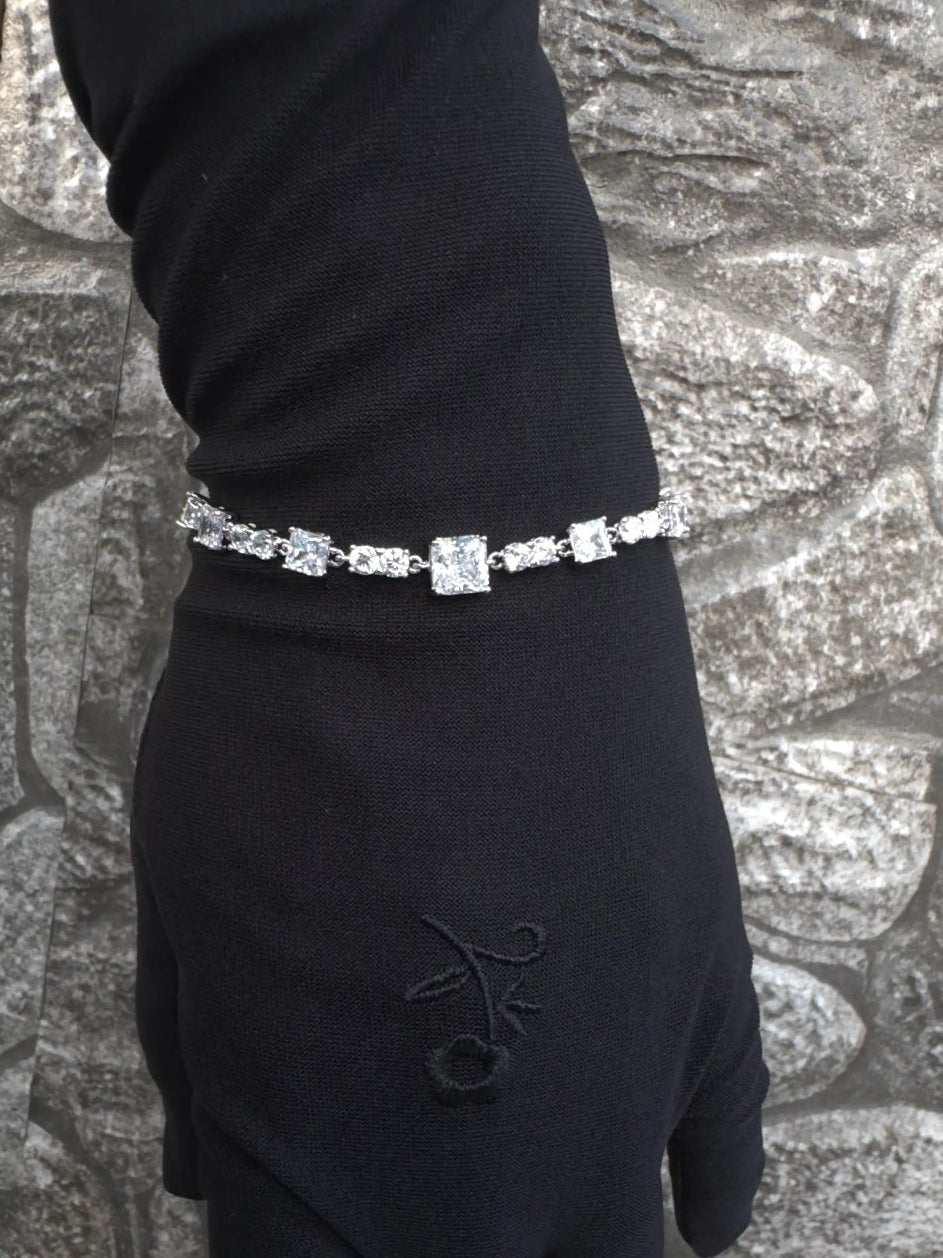 Hand wearing a black glove with a diamond bracelet against a textured stone wall.