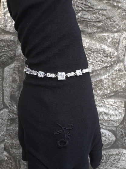 Hand wearing a black glove with a diamond bracelet against a textured stone wall.