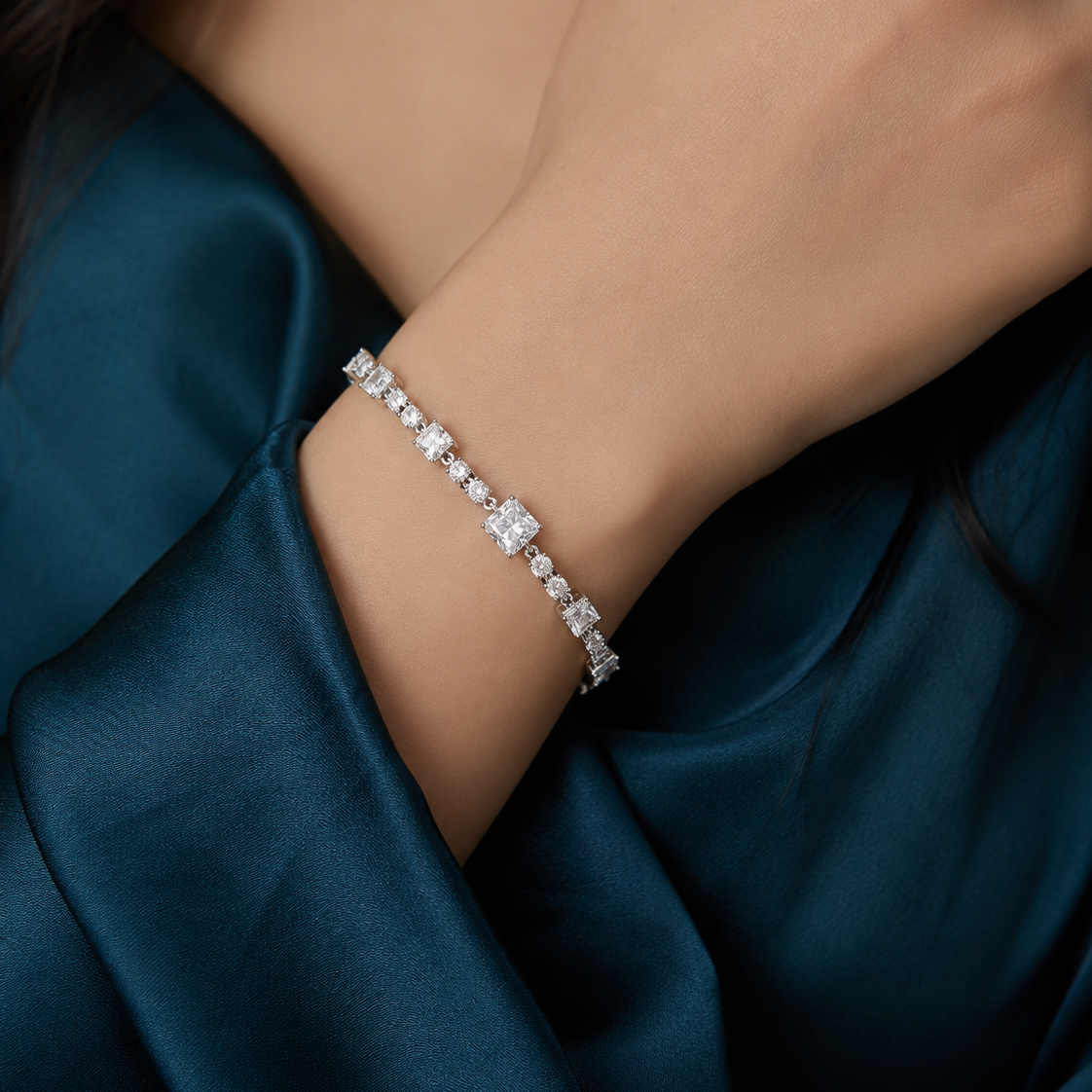 A close-up of a delicate silver tennis bracelet with clear stones worn on a woman's wrist, set against a dark teal satin fabric background.