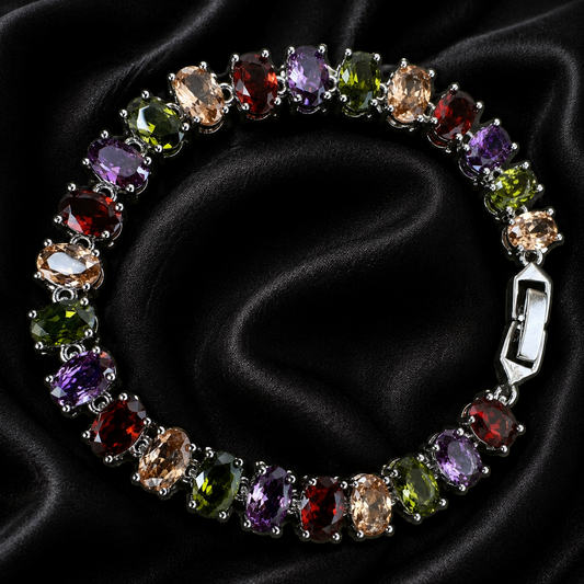 A multi-colored gemstone tennis bracelet with oval-cut stones in purple, green, red, and peach tones, set in silver and displayed on a black satin background.