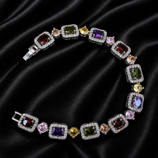 A multi-color gemstone bracelet arranged in a circle on a black satin fabric background. The bracelet features square and round-cut colored gemstones in silver-toned settings with clear accent stones.