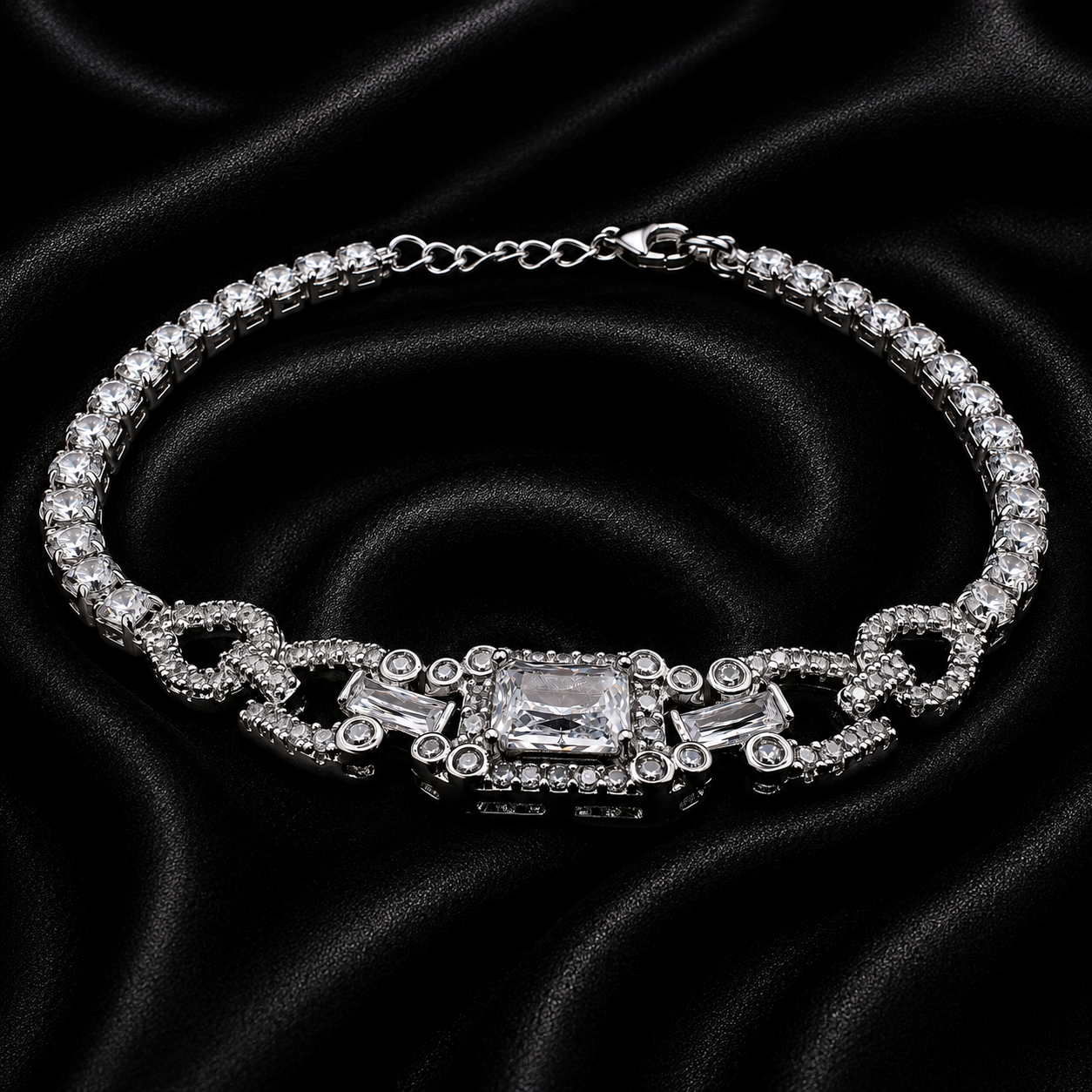 A silver-tone tennis bracelet with clear crystals, featuring a central rectangular crystal and decorative links, displayed on a black satin fabric background.
