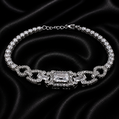 A silver-tone tennis bracelet with clear crystals, featuring a central rectangular crystal and decorative links, displayed on a black satin fabric background.