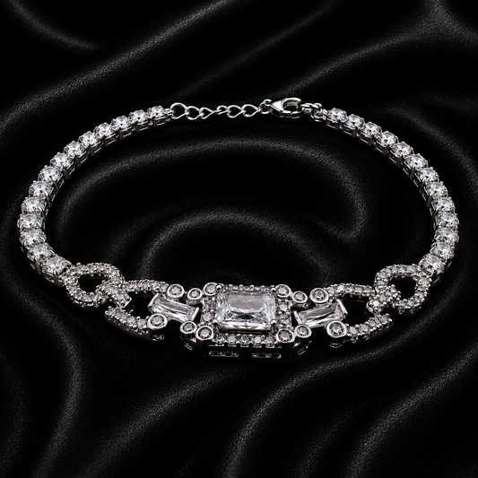 A silver-tone tennis bracelet with clear crystals, featuring a central rectangular crystal and decorative links, displayed on a black satin fabric background.