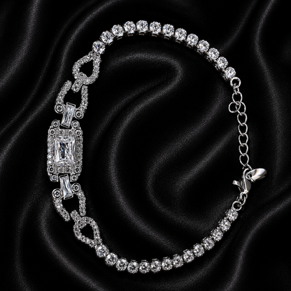 A silver-tone tennis bracelet with clear gemstones, featuring a central rectangular emerald-cut stone and decorative links, displayed on a black satin fabric background.